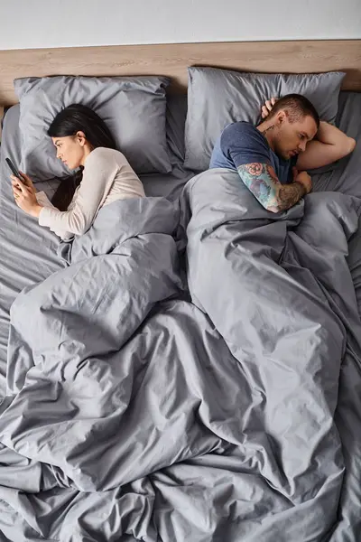 Giovane uomo tatuato vicino moglie asiatica guardando il suo smartphone in camera da letto, fraintendimento, vista dall'alto — Foto stock