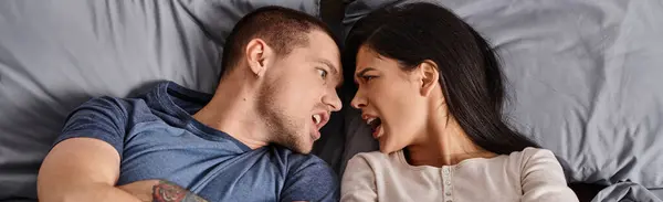 Top view of irritated multiethnic couple lying down with folded arms and quarreling on bed, banner — Stock Photo