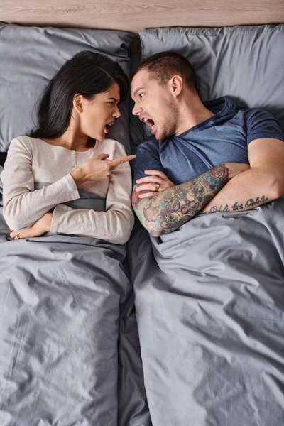 Top view of angry multicultural couple lying down on bed screaming and quarreling at each other — Stock Photo