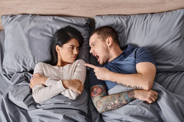 Top view of irritated interracial couple lying down on bed shouting and quarreling at each other — Stock Photo