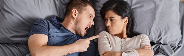 Angry multicultural couple lying down on bed shouting and quarreling at each other, top view, banner — Stock Photo