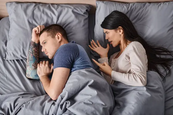 Top view of angry asian woman quarreling at tattooed young husband lying down on bed, divorce — Stock Photo