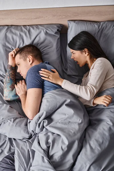 Top view of irritated asian woman quarreling at tattooed young husband lying down on bed, divorce — Stock Photo