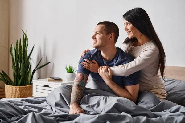Cheerful asian woman hugging young tattooed man looking away in bedroom at home, joy and happiness — Stock Photo
