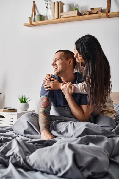 Mujer asiática encantada abrazando joven hombre tatuado mirando hacia otro lado en el dormitorio en casa, alegría y felicidad - foto de stock