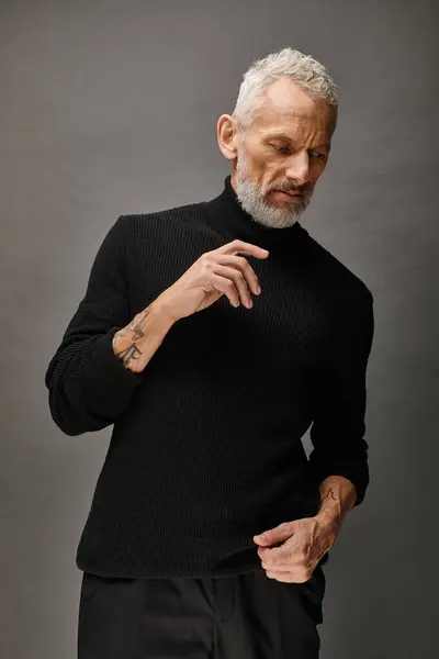 Atractivo hombre maduro de moda en moda cuello alto posando y mirando hacia otro lado en el fondo gris - foto de stock
