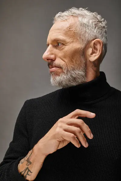 Guapo hombre maduro de moda en elegante cuello alto negro posando y mirando hacia otro lado en el fondo gris - foto de stock