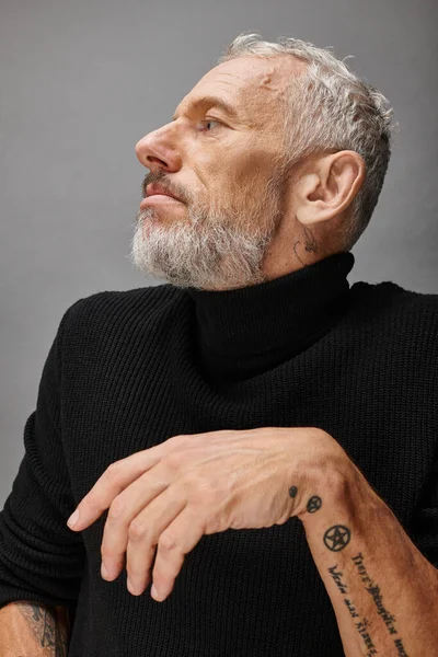 Guapo maduro hombre fashionista con barba gris en elegante cuello alto negro posando y mirando hacia otro lado - foto de stock