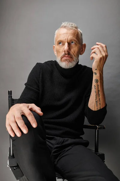 Handsome gray bearded mature man in fashionable turtleneck sitting on folding chair and looking away — Stock Photo