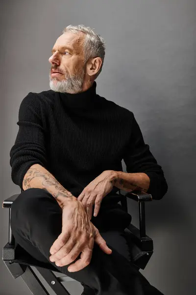 Bonito cinza barbudo maduro homem na moda gola alta sentado na cadeira dobrável e olhando para longe — Fotografia de Stock