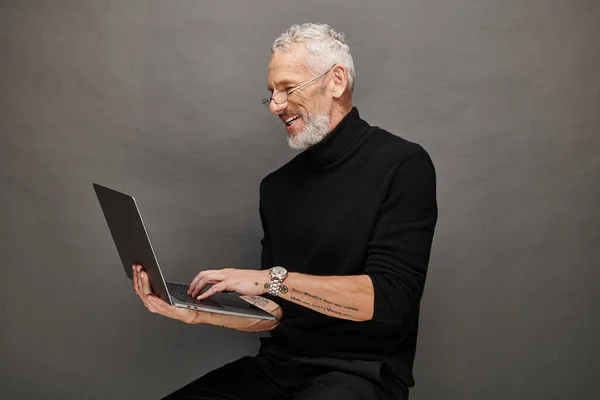 Cheerful handsome bearded mature man with glasses sitting on chair with laptop and smiling happily — Stock Photo