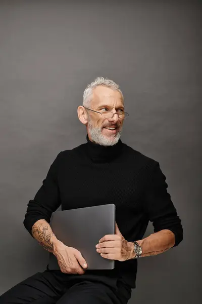 Cheerful handsome bearded mature man with glasses sitting on chair with laptop and smiling happily — Stock Photo