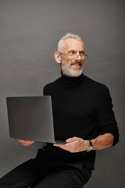 Joyous attractive bearded mature man with glasses sitting on chair with laptop and smiling happily — Stock Photo