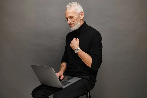 Gut aussehender bärtiger älterer Mann in schwarzem Rollkragen sitzt auf einem Stuhl und schaut auf seinen Laptop — Stockfoto