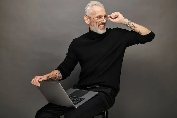 Alegre atraente barbudo maduro homem com óculos sentado na cadeira com laptop e sorrindo alegremente — Fotografia de Stock