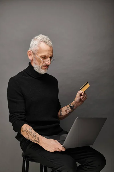 Good looking bearded mature man with glasses in turtleneck holding credit card and looking at laptop — Stock Photo