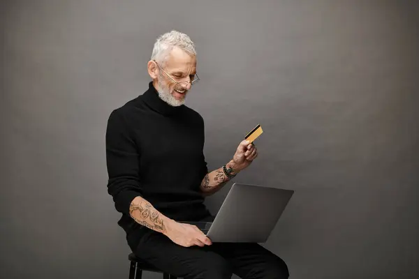Веселый привлекательный зрелый мужчина в очках в водолазке с кредиткой и глядя на ноутбук — стоковое фото
