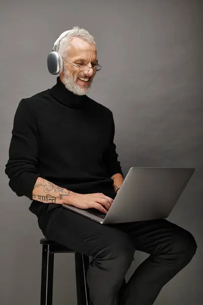 Homem bonito alegre com óculos e fones de ouvido sentado na cadeira com laptop e sorrindo sinceramente — Fotografia de Stock