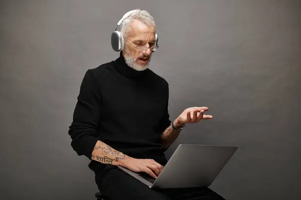 Good looking bearded man in stylish black turtleneck with headphones sitting and having video call — Stock Photo