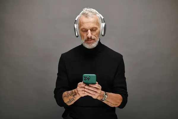 Good looking mature male model with beard and headphones in black turtleneck looking at phone — Stock Photo