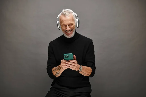 Joyous attractive mature male model with beard and headphones in black turtleneck looking at phone — Stock Photo