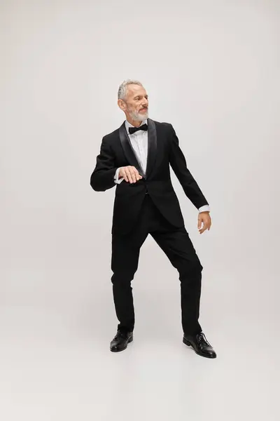 Modelo masculino maduro bien vestido con pajarita en esmoquin negro chic bailando activamente sobre fondo gris - foto de stock