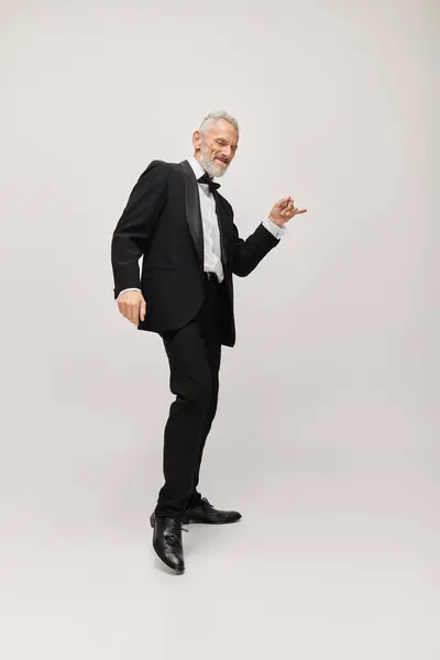 Joyful good looking mature man in elegant suit with bow tie dancing happily on gray backdrop — Stock Photo