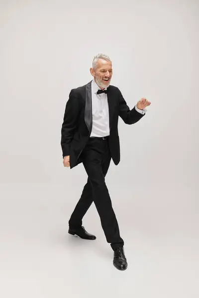 Joyous good looking mature man in elegant suit with bow tie dancing happily on gray backdrop — Stock Photo