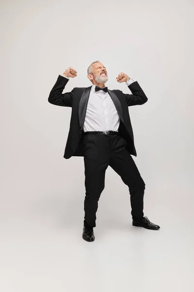 Joyous good looking mature man in elegant suit with bow tie dancing happily on gray backdrop — Stock Photo