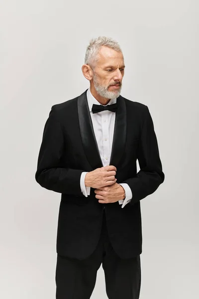 Гарний виглядає бородатий зрілий чоловік в елегантному чорному смокінгу з краваткою позує і дивиться геть — стокове фото