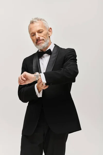 Hombre barbudo maduro atractivo con corbata de lazo en elegante esmoquin posando y mirando reloj de pulsera - foto de stock