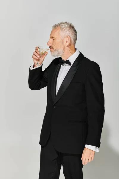 Homem maduro bonito em smoking elegante com barba cinza e gravata borboleta bebendo delicioso martini — Fotografia de Stock