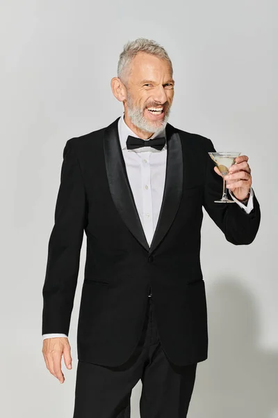 Homem maduro bonito alegre em smoking chique com barba cinza sorrindo e segurando martini com azeitonas — Fotografia de Stock