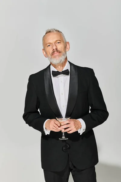 Schöne bärtige reife männliche Modell im eleganten Smoking hält Martini-Glas und schaut in die Kamera — Stockfoto