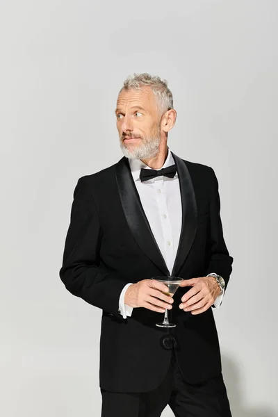 Homem maduro de boa aparência em smoking elegante com barba segurando martini com azeitonas e olhando para longe — Fotografia de Stock