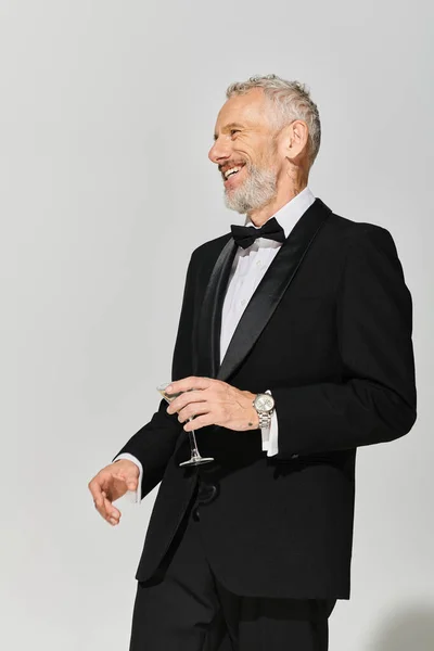 Happy elegant mature man with beard in debonair tuxedo holding martini and smiling cheerfully — Stock Photo