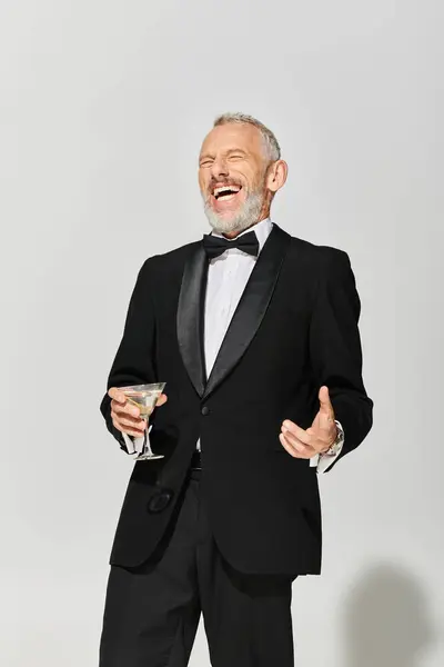 Feliz elegante maduro homem com barba em debonair smoking segurando martini e sorrindo alegremente — Fotografia de Stock