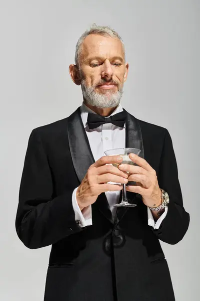 Attraktiver bärtiger älterer Mann im eleganten Smoking mit Fliege, der ein Glas Martini mit Oliven hält — Stockfoto