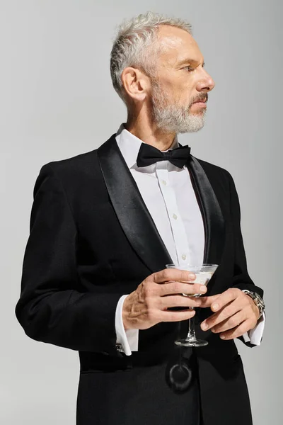 Bonito barbudo maduro homem no elegante smoking com laço gravata segurando martini e olhando para longe — Fotografia de Stock