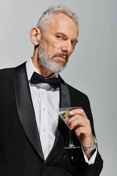 Gut aussehender bärtiger älterer Mann im eleganten Smoking hält ein Glas Martini in der Hand und schaut in die Kamera — Stockfoto