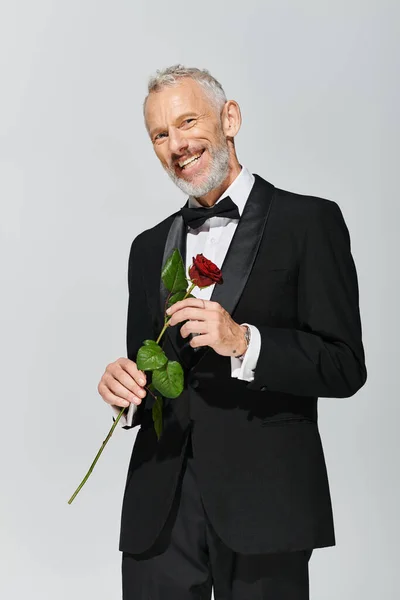 Привабливий веселий зрілий чоловік з бородою в елегантному смокінгу тримає червону троянду і посміхається на камеру — стокове фото