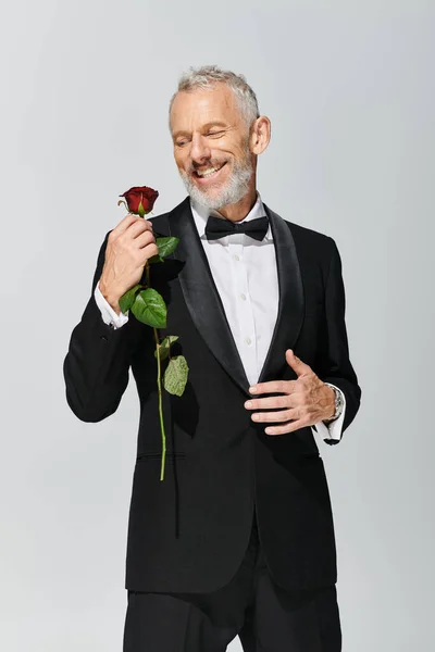 Atractivo hombre maduro alegre con barba en elegante esmoquin sosteniendo rosa roja y sonriendo felizmente - foto de stock