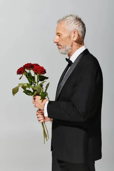 Элегантный красивый зрелый мужчина в модном черном смокинге с букетом красных роз в руках — стоковое фото