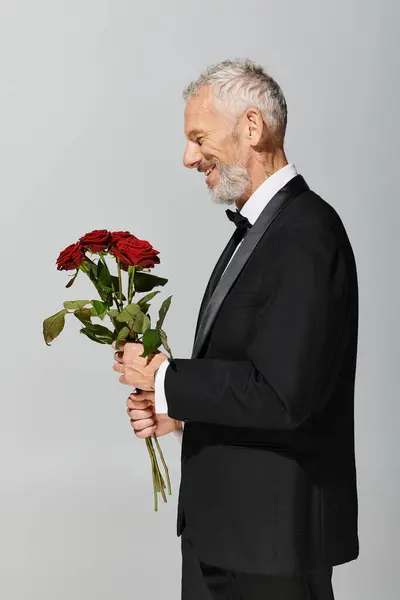 Cheerful good looking mature man in voguish black tuxedo holding red roses bouquet in hands — Stock Photo