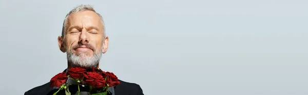 Freudig gut aussehender reifer Mann im schicken Smoking mit roten Rosen Strauß und Lächeln mit geschlossenen Augen — Stockfoto