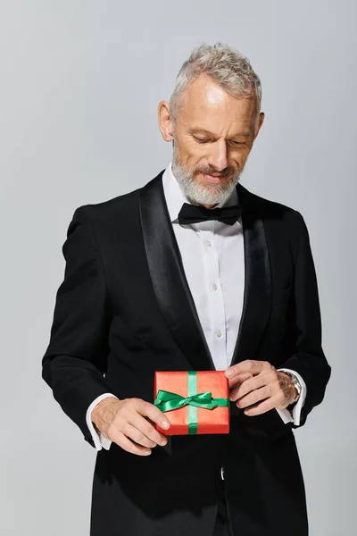 Good looking elegant mature male model with beard and bow tie in tuxedo looking at gift in his hands — Stock Photo