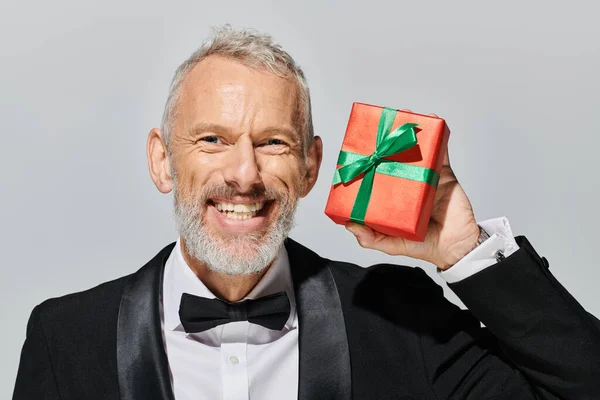 Cheerful good looking voguish mature man with beard in tuxedo holding gift and smiling at camera — Stock Photo