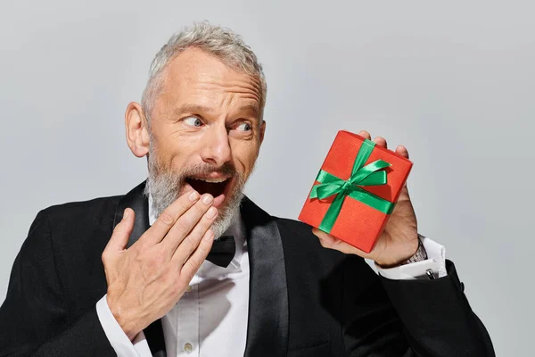 Cheerful surprised mature man with beard in elegant black tuxedo looking at present in his hand — Stock Photo