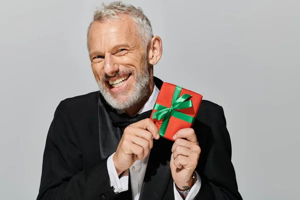 Joyous good looking debonair mature man with beard in tuxedo holding gift and smiling at camera — Stock Photo