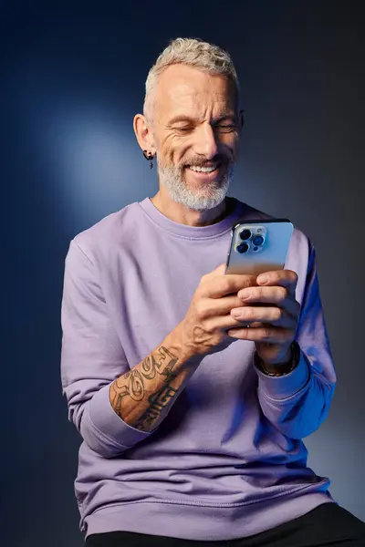 Joyous good looking mature man in purple sweatshirt with gray beard looking at his smartphone — Stock Photo